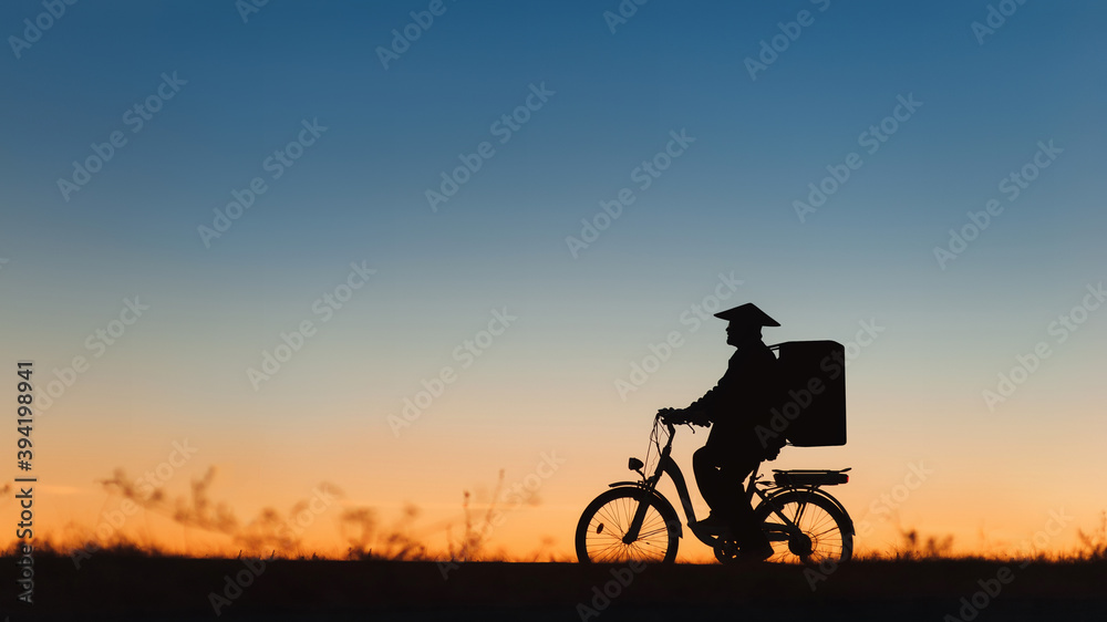 Obraz premium An elderly Asian man in a Vietnamese bamboo hat with a basket on his back rides an electric bike. The concept of retirement age and the delivery of goods. Silhouette against the sunset.