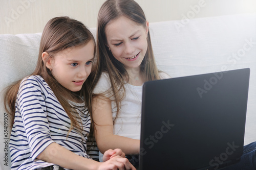 Two schoolgirls sitting at home on the couch emotionally looking at a laptop.