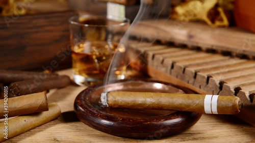 Burning Cigar in an Ashtray with Whisky and Tobacco Leaves on wooden Background