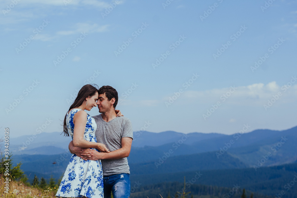 Stylish young couple in the summer mountains. A guy and a girl hug together on a background of a forest and mountain peaks at midday. Stylish young couple in the summer mountains. A guy and a girl hug together on a background of a forest and mountain peaks at midday.