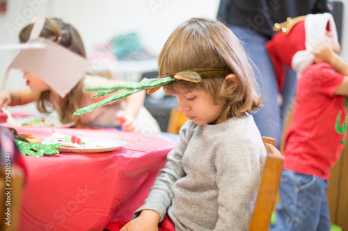 Small child male gender boy wearing green reindeer headband celebrating christmas holiday season sitting at a table with red tablecloth at school looking down surrounded by classmates