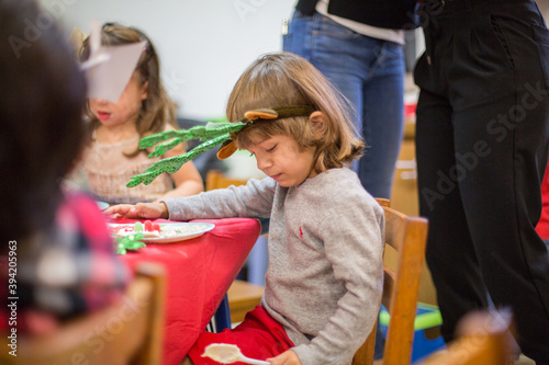 Small child male gender boy wearing green reindeer headband celebrating christmas holiday season sitting at a table with red tablecloth at school looking down surrounded by classmates
