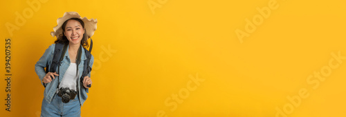 panoramic wide banner. asian woman hipster traveler with camera and hat in casual clothing happy and smiling over copy space yellow color background, summer holiday, backpacker travel concept
