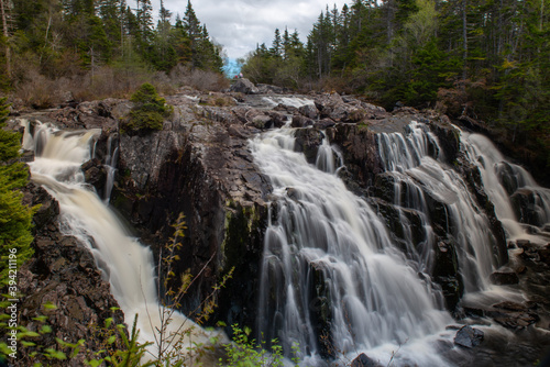 Multiple waterfalls of freshwater overflowing over a rocky wilderness area. The falls have trees, clouds, and a blue sky in the background. The drop is steep, allowing the river water to cascade. 