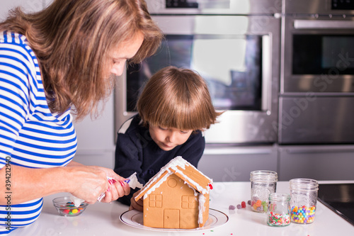 Boy child with grandmother or mother decorating a Christmas gingerbread house during the holidays using icing and white frosting as well as colorful sprinkles, candy cane, and gummy candy in kitchen