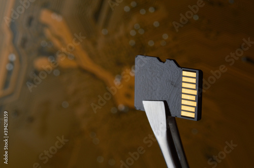 Close-up of a Micro SD memory card against the background of a microcircuit, clamped with tweezers.