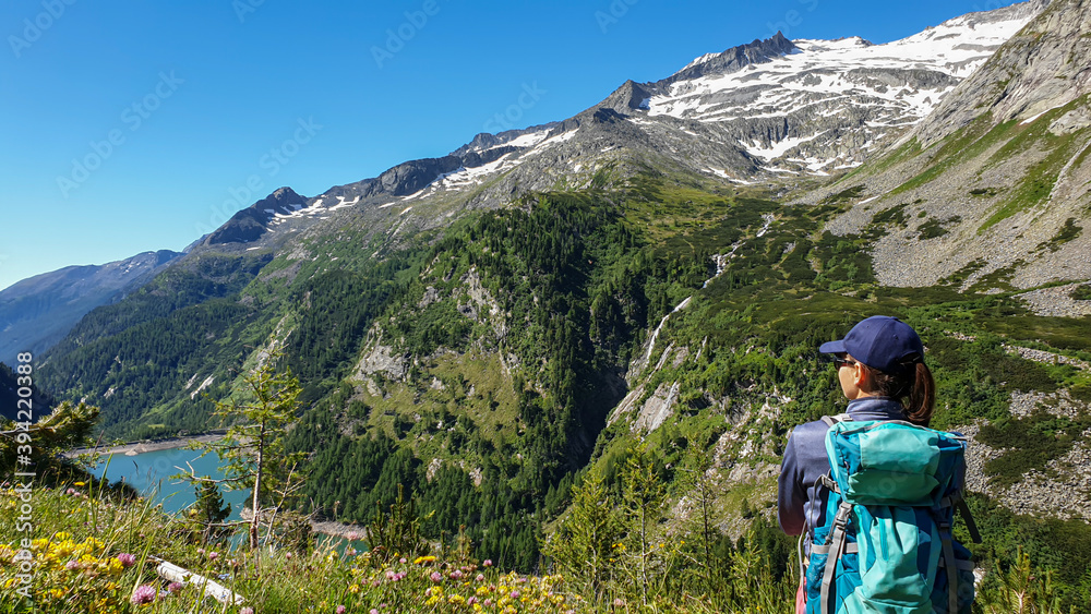 A woman sitting at the side of a mountain and enjoying the view on a lake below at the K?lnbreinsperre in Austria. High peaks are covered with snow. The meadows blossoming with wild flowers. Calmness