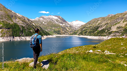 Man standing at the side of a lake at K?lnbreinsperre dam in Austria. The lake has navy blue color. High Alps around. There is a glacier in the back. The meadows blossoming with wild flowers. Calmness photo