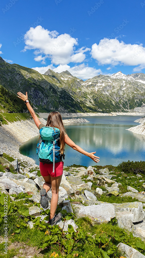 A woman with hiking backpack standing at the side of an artificial lake at a dam in Austria. The lake has navy blue color. High Alps around. There is a glacier in the back. Adventure and discovering