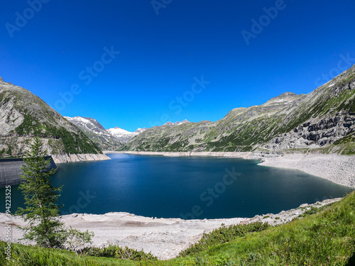 A panoramic view on an artificial  dam lake stretching over a vast territory around Alps in Austria. Lake is shining with navy blue color. In the back there are a few glaciers. Controlling the nature
