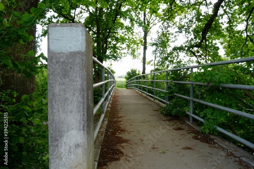 A narrow bridge with metal railings and concrete pylon surrounded by lush green vegetation diminishing in the perspective of a sun flooded land. Photo with copy space and potentially symbolic meaning.