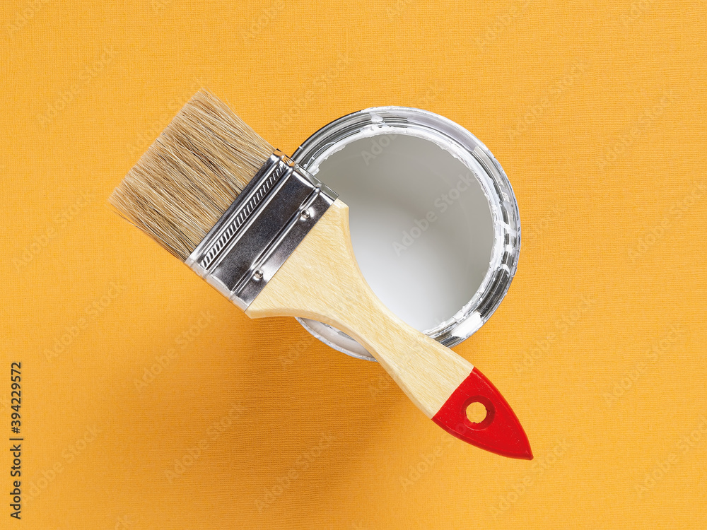New natural bristle brush with wooden handle on the edge of an open white paint can over bright yellow textured background. Construction painting work, repair and redecorate concept. New natural bristle brush with wooden handle on the edge of an open white paint can over bright yellow textured background. Construction painting work, repair and redecorate concept.