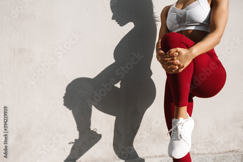 Cropped image of a fitness woman stretching her legs before jogging workout outdoors. Fit female runner doing stretches, warming-up for exercises, shadow on concrete wall
