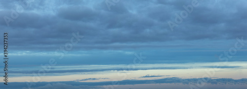 Background of gray cumulus clouds with blue and yellow stripes from the rising sun at dawn hour. Long panoramic banner