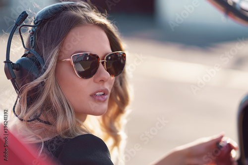 Young fashionable woman pilot in headset ready to fly in small red airplane. Beautiful life, aristocratic lady in black dress in blonde wavy hair.