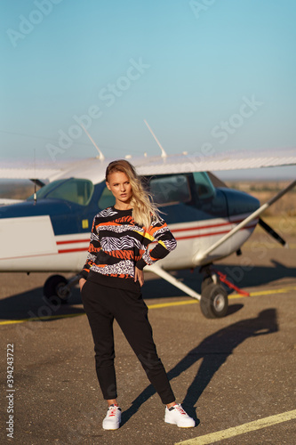 Young woman model with a modern haircut and fashionable sunglasses posing near a red plane wearing trendy casual outfit © diignat