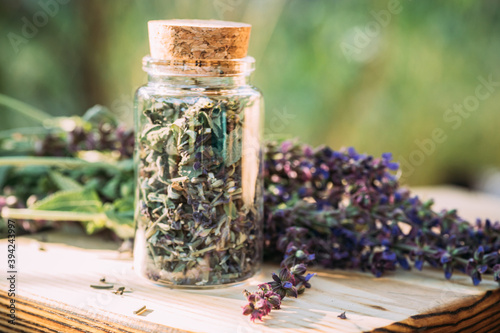 Transparent jar with cortical cork with dry grass meadow sage purple fresh flowers near mortar with cooked dried herb. 