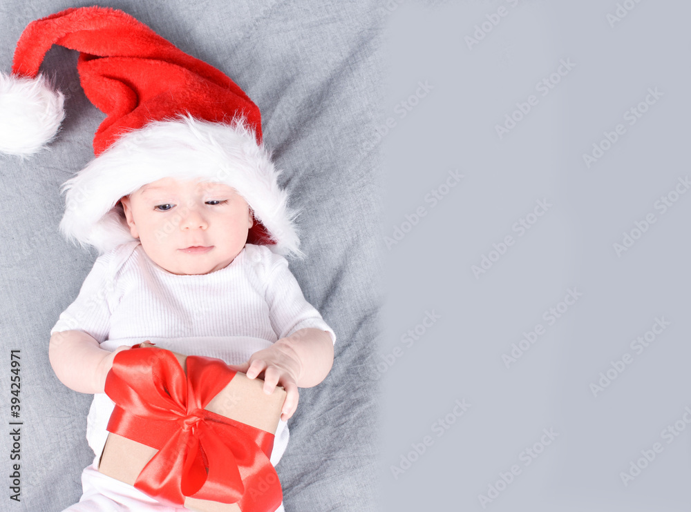 Christmas mood. Cute four month old baby in a red Santa Claus hat lying on back holding/looking at gift on grey background. Interested look. Copy space. Christmas card. Christmas mood. Cute four month old baby in a red Santa Claus hat lying on back holding/looking at gift on grey background. Interested look. Copy space. Christmas card.