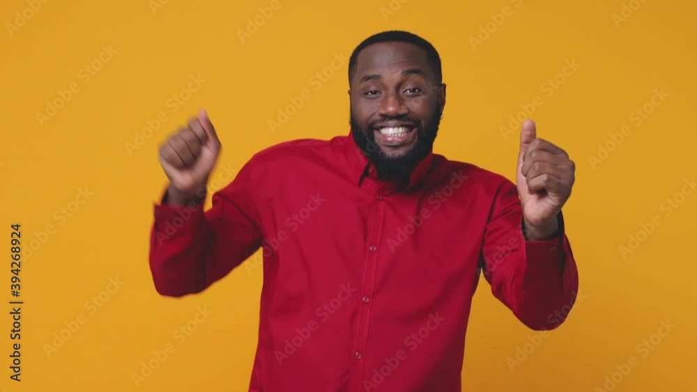 Smiling cheerful excited young bearded african american man 20s wearing red shirt isolated on bright yellow color background studio. People lifestyle concept. Looking camera dancing clenching fists