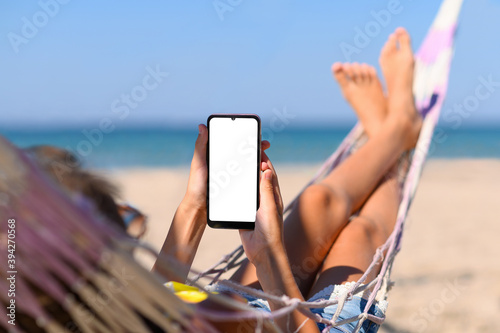 Smartphone with a blank white display in the hands of a girl against the background of the sea. The girl lies on a hammock on the beach with a phone in her hands.