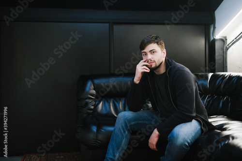 Adult man with a beard and casual clothes sits on a leather sofa in a dark room with a black wall, posing for a camera with a serious face. Man relaxing sitting on black couch, looking into camera. © bodnarphoto
