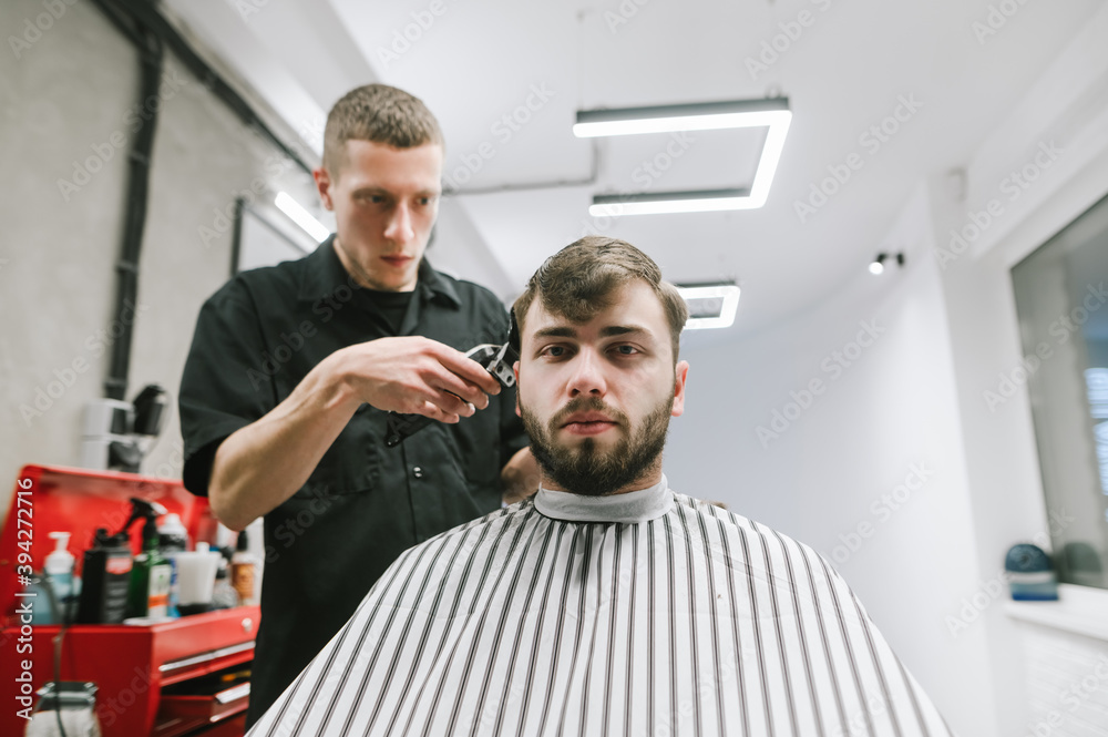 Φωτογραφία Adult bearded man in a dressing room sits in a barbershop chair, cuts hair at a hairdresser, looks in camera with a serious face
