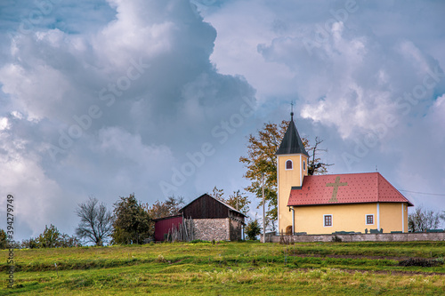 Landscape view of small church and cross sign on roof, near by rural house . Green meadow at foot. Natural park, Samoborsko gorje, Croatia.