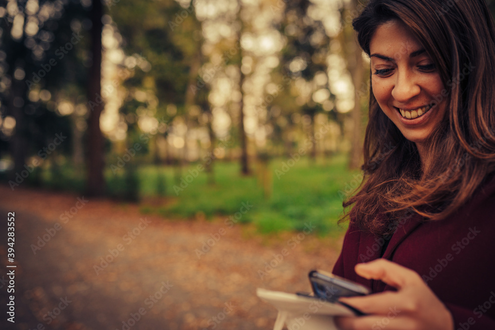 young woman smiling while using her smartphone in the middle of a park with autumn colors young woman smiling while using her smartphone in the middle of a park with autumn colors
