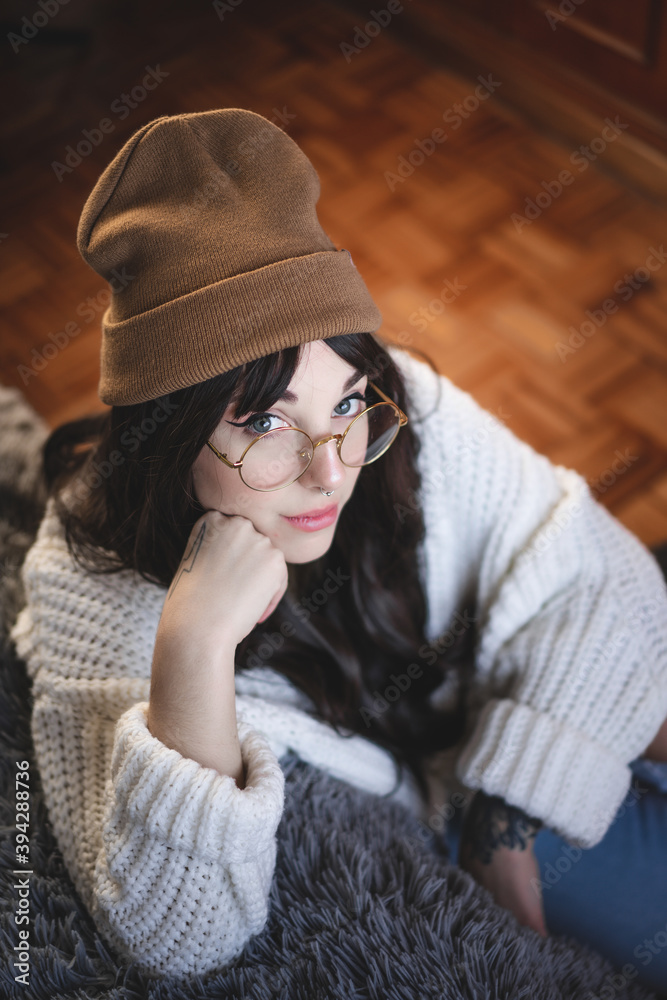 Foto de Mujer caucásica joven, guapa, con gorro de lana marrón, sueter de  lana, gafas redondas, pelo moreno, apoyada en una cama, con fondo de  madera. Otoño. do Stock