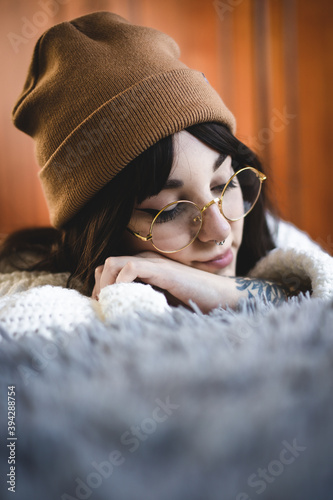 Mujer caucásica joven, guapa, con gorro de lana marrón, sueter de lana, gafas  redondas, pelo moreno, apoyada en una cama, con fondo de madera. Otoño.  Stock Photo