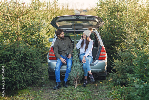 Couple in love loading freshly cut down christmas tree into a trunk of their car. Young family preparing to holidays. © gorynvd
