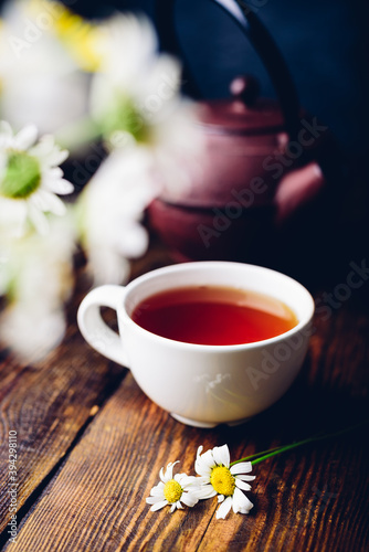 Cup of red tea, two chamomile flowers and kettle on wooden table. Blurred bouquet on foreground