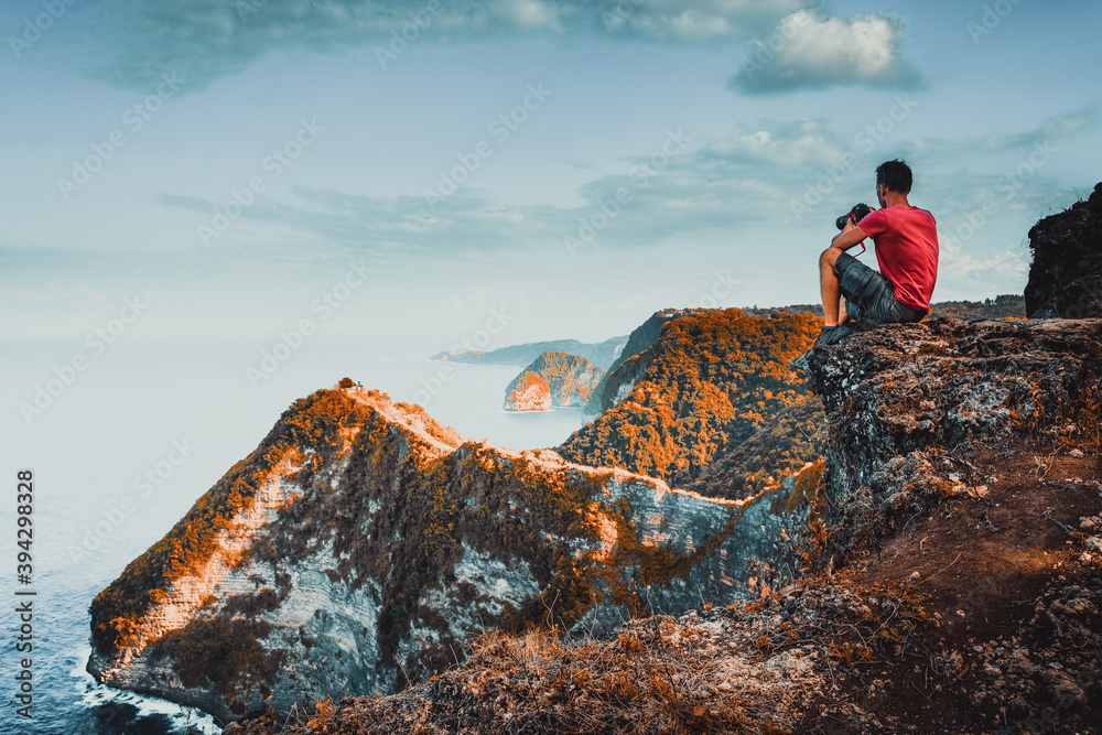 A traveler photographer with a camera in hand sitting on top of a mountain and enjoys an amazing nature landscape at sunrise. Active lifestyle and Travel concept.