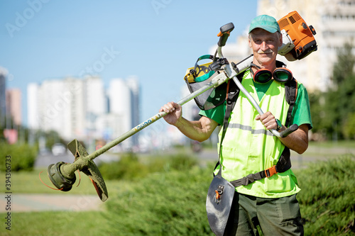 Municipal gardener landscaper senior man worker with gas grass trimmer equipment on sunlight city background