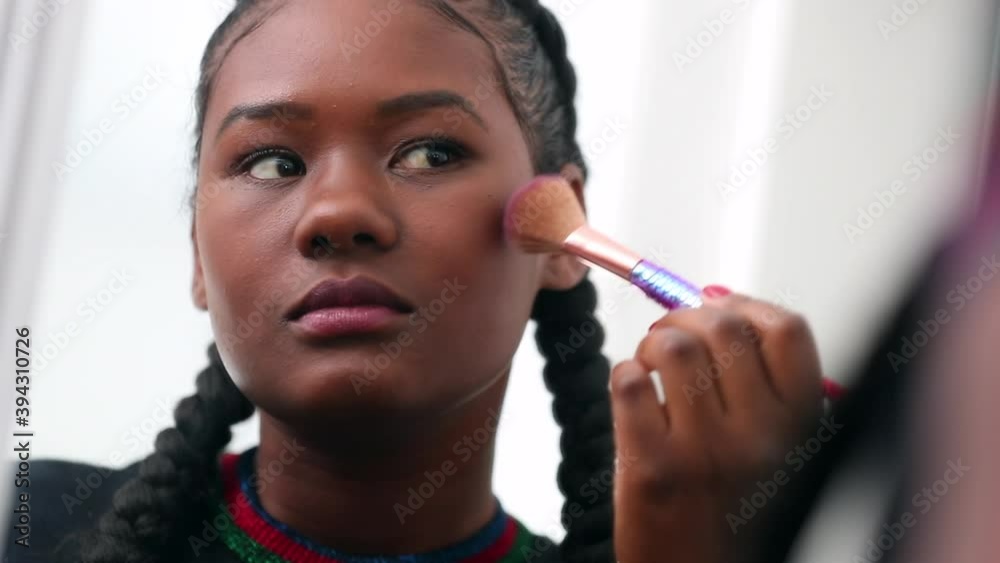 Black girl applies make-up powder. African young woman looking at mirror