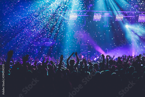 A crowded concert hall with scene stage lights, rock show performance, with people silhouette, colourful confetti explosion fired on dance floor air during a concert festival