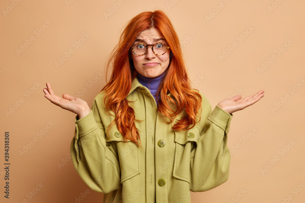 Human perception concept. Doubtful confused hesitant ginger woman raises palms and shrugs shoulders questioned faces difficult choice wears fashionable coat isolated over brown studio background