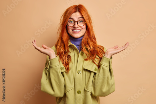 Unaware confused cheerful young European woman with ginger hair raises palms has doubtful expression cannot give answer immediately wears green jacket isolated over brown background. Who cares