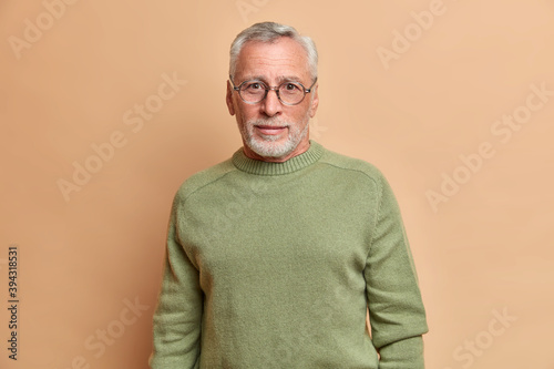 Handsome bearded European man with curious gaze wears spectacles and basic jumper looks directly at camera poses against beige background. Self confident successful senior entrepreneur indoor © wayhome.studio 