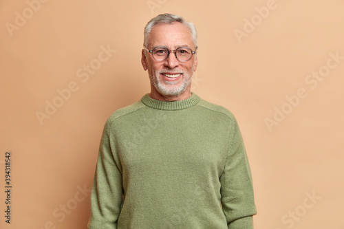 Half length shot of cheerful senior man smiles happily with white teeth wears optical glasses and sweater isolated over brown background. Happy grandfather glad grandchildren came to visit him photo