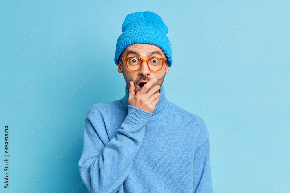 Shocked surprised man covers mouth and gazes at camera with stupefied expression hears unbelievable rumors wears blue hat and turtleneck poses indoor. Emotional hipster guy impressed by something Shocked surprised man covers mouth and gazes at camera with stupefied expression hears unbelievable rumors wears blue hat and turtleneck poses indoor. Emotional hipster guy impressed by something