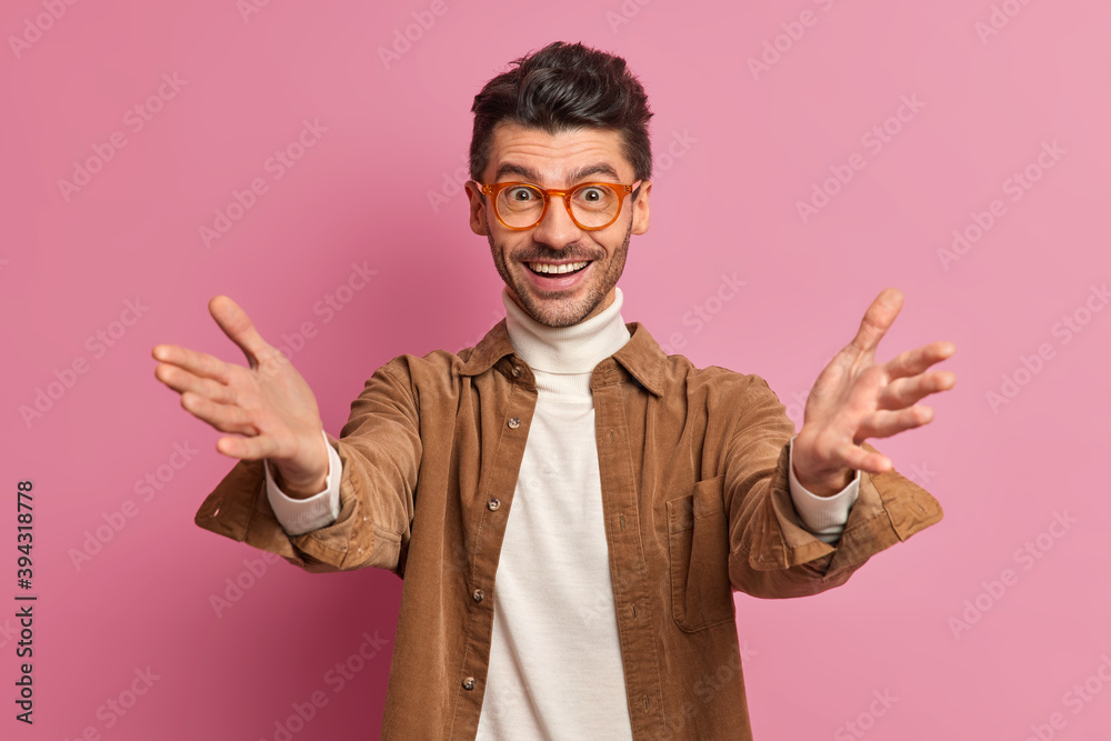 Positive friendly European man raises hands as meets someone pulls arms in welcome gesture wants to embrace friend smiles gladfully wears stylish shirt and spectacles isolated over pink background Positive friendly European man raises hands as meets someone pulls arms in welcome gesture wants to embrace friend smiles gladfully wears stylish shirt and spectacles isolated over pink background