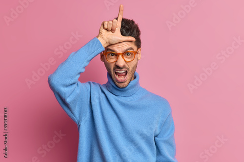 Annoyed adult man makes looser gesture wears spectacles and blue turtleneck has failure or unlucky day as lost his job upset about dismissal makes L finger sign over forehead isolated on pink wall photo