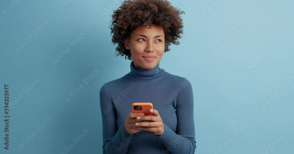 Cheerful curly haired young female in casual turtleneck types message in hurry uses smartphone for online communication enjoys browsing social media poses against blue background reads received sms Cheerful curly haired young female in casual turtleneck types message in hurry uses smartphone for online communication enjoys browsing social media poses against blue background reads received sms