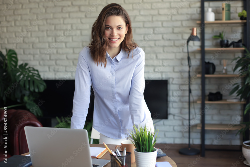 Confident young businesswoman with a friendly smile standing behind her desk in a home office looking at the camera. Confident young businesswoman with a friendly smile standing behind her desk in a home office looking at the camera.