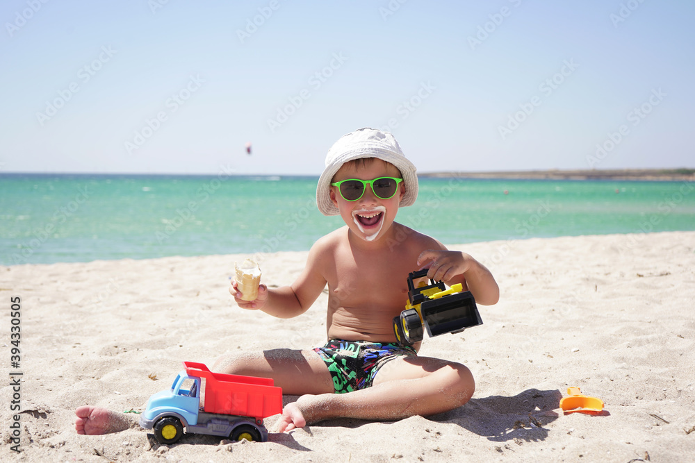 on a Sunny hot summer day, a boy plays with sand and toys, cars on the beach. a child eats ice cream while sitting on a sandy beach with an azure sea background. on a Sunny hot summer day, a boy plays with sand and toys, cars on the beach. a child eats ice cream while sitting on a sandy beach with an azure sea background.