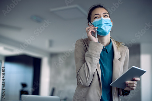 Happy businesswoman with face mask talking on the phone while using touchpad in the office.
