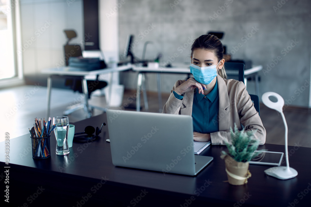 Female entrepreneur with face mask reading an e-mail on laptop while working in the office.