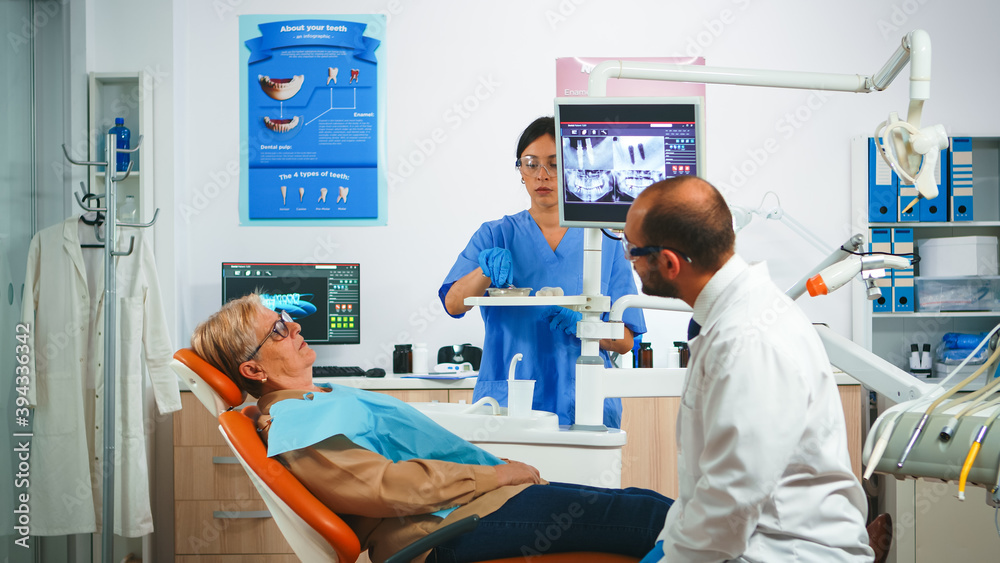 Obraz premium Man dentistry doctor pointing on digital screen showing dental implants. Dentist and nurse working together in modern stomatological clinic, explaining radiography of teeth on monitor