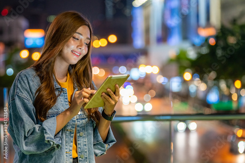 Smiling young Asian woman using digital tablet for social media or online shopping in the city at night with illuminated city night lights background. Happy pretty girl enjoy with city nightlife.